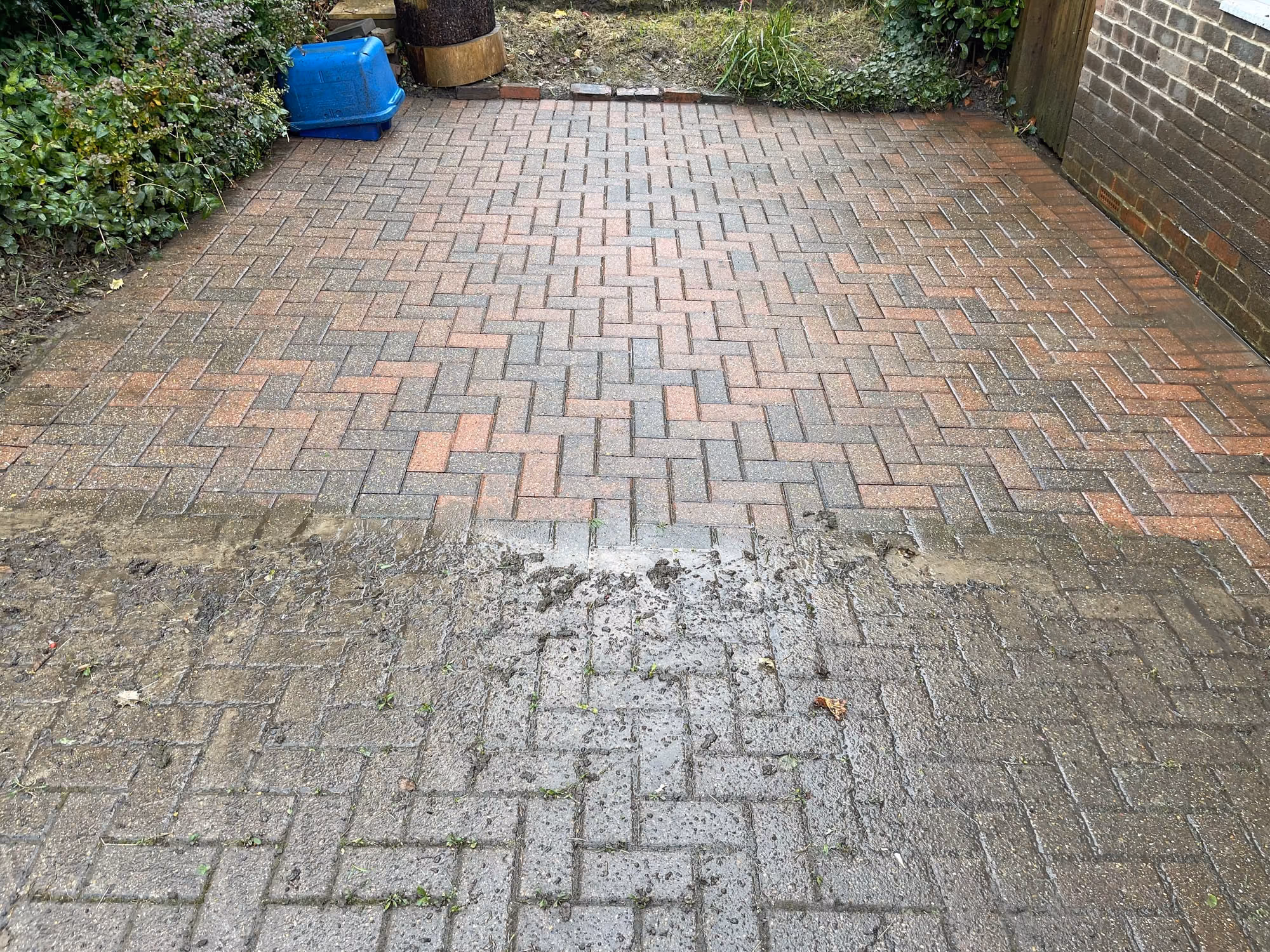 A block paving driveway halfway through cleaning, with a cleaned strip cut across the dirty grey paving showing the red and buff colour underneath