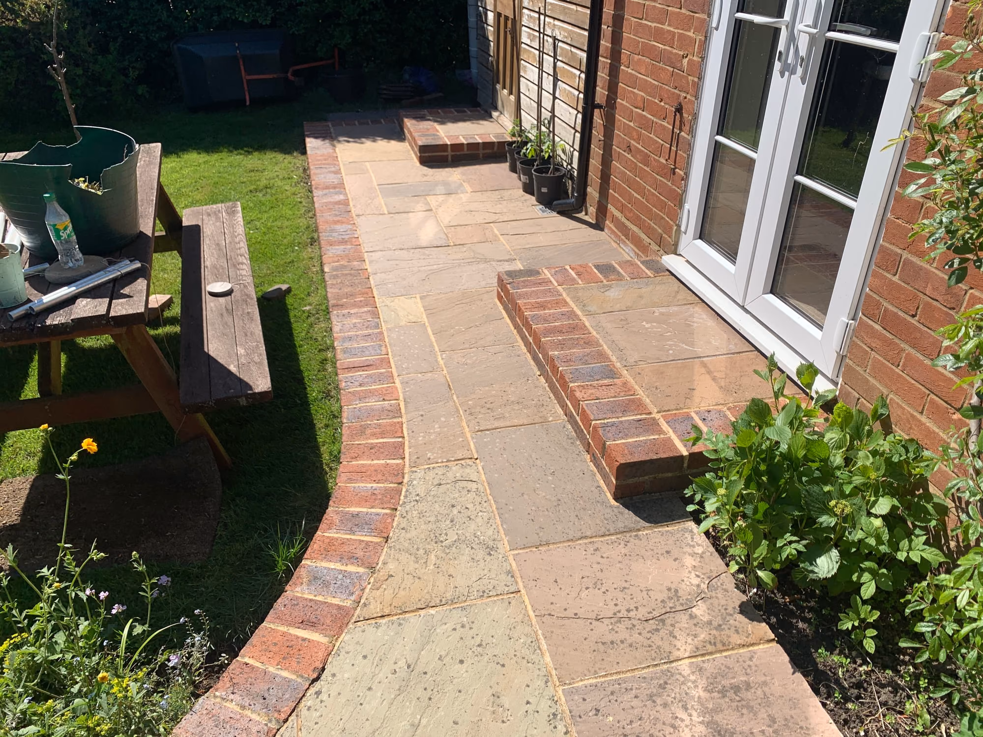Indian sandstone paving with a brick step and brick edging outside a pair of white French doors, cleaned, with a lawn and garden table beyond