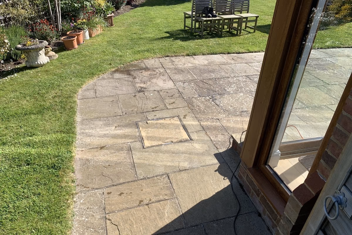 Indian sandstone patio beside a lawn, the slabs flattened to a dull grey by a film of algae, with moss showing along the joints