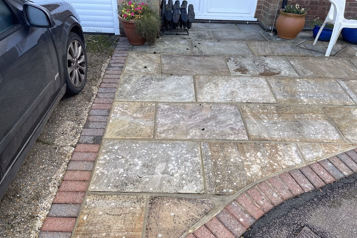 Sandstone patio next to a parked car, the slabs stained dark grey with algae and lichen spotting, edged with red brick